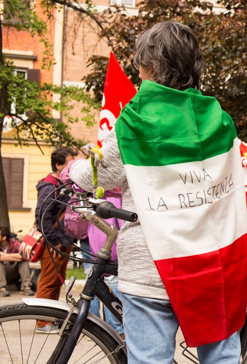 una persona di spalle indossa una bandiera italiana con la scritta "VIVA LA RESISTENZA" come un mantello a una manifestazione per il 25 aprile.